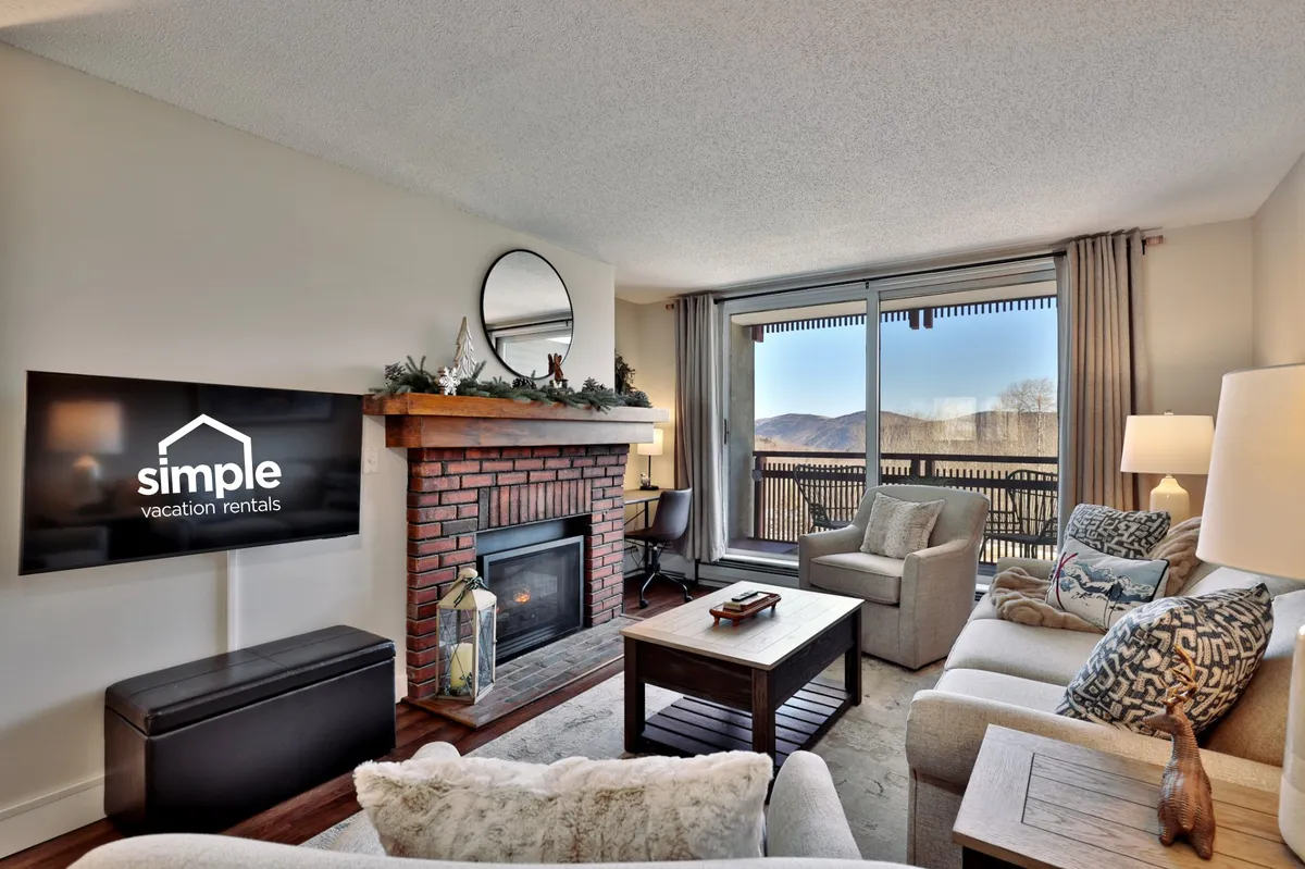 A warm, comfortable living room featuring a gas fireplace as a central focal point. The space includes a plush sofa, large windows that fill the room with natural light, and a television for entertainment. The windows offer an inviting glimpse of the outdoors, enhancing the sense of relaxation and connection to the mountain setting.