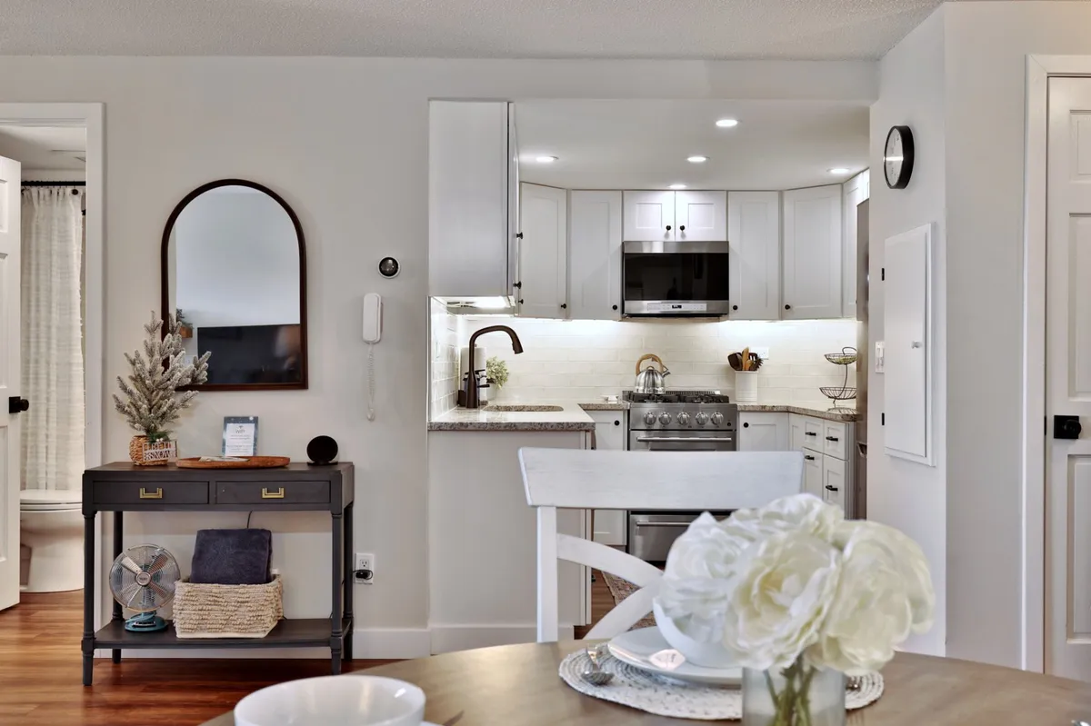 A bright dining area adjacent to the kitchen, featuring a dining table adorned with fresh flowers and a glimpse of the kitchen area and a wall mirror, creating a sense of openness and connection between spaces.