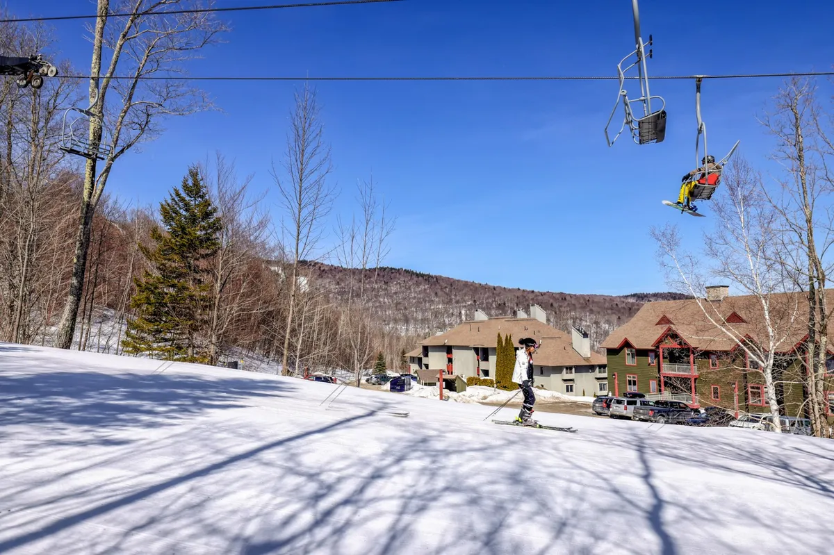A dynamic exterior winter scene featuring a ski lift in motion, a skier traversing the snow-covered terrain, and a backdrop of majestic mountains. Residential buildings are visible in the distance, highlighting both the adventure of the slopes and the close proximity of accommodations.