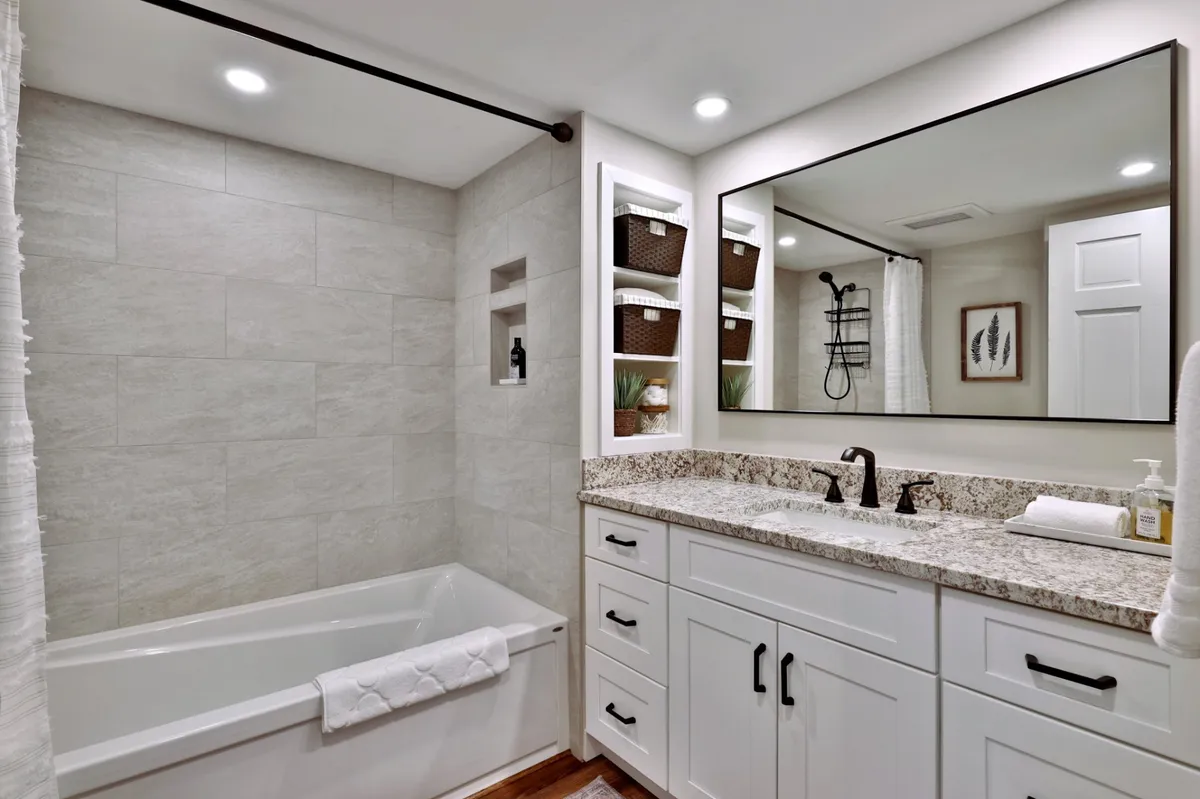 A modern bathroom is displayed, featuring a tranquil bathtub, a sleek vanity with sink, large mirror, and open storage shelves. The scene is bright, organized, and equipped with fresh towels and essentials, embodying a spa-like retreat.