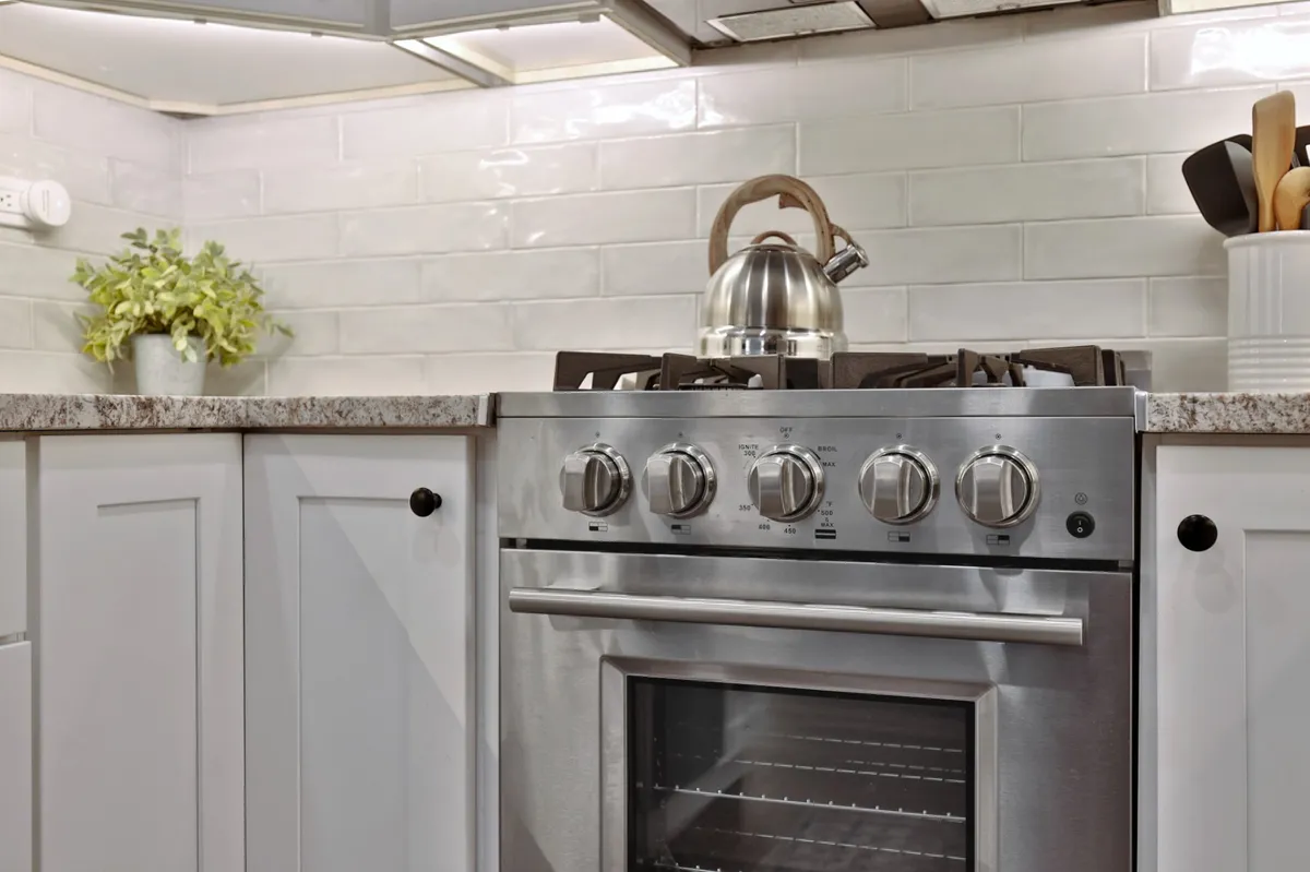 A contemporary kitchen view focusing on a stainless steel oven, granite countertops, a kettle ready for morning coffee, and white cabinetry accented by a pop of green from a potted plant. The environment is functional yet welcoming, ideal for both cooking and entertaining.