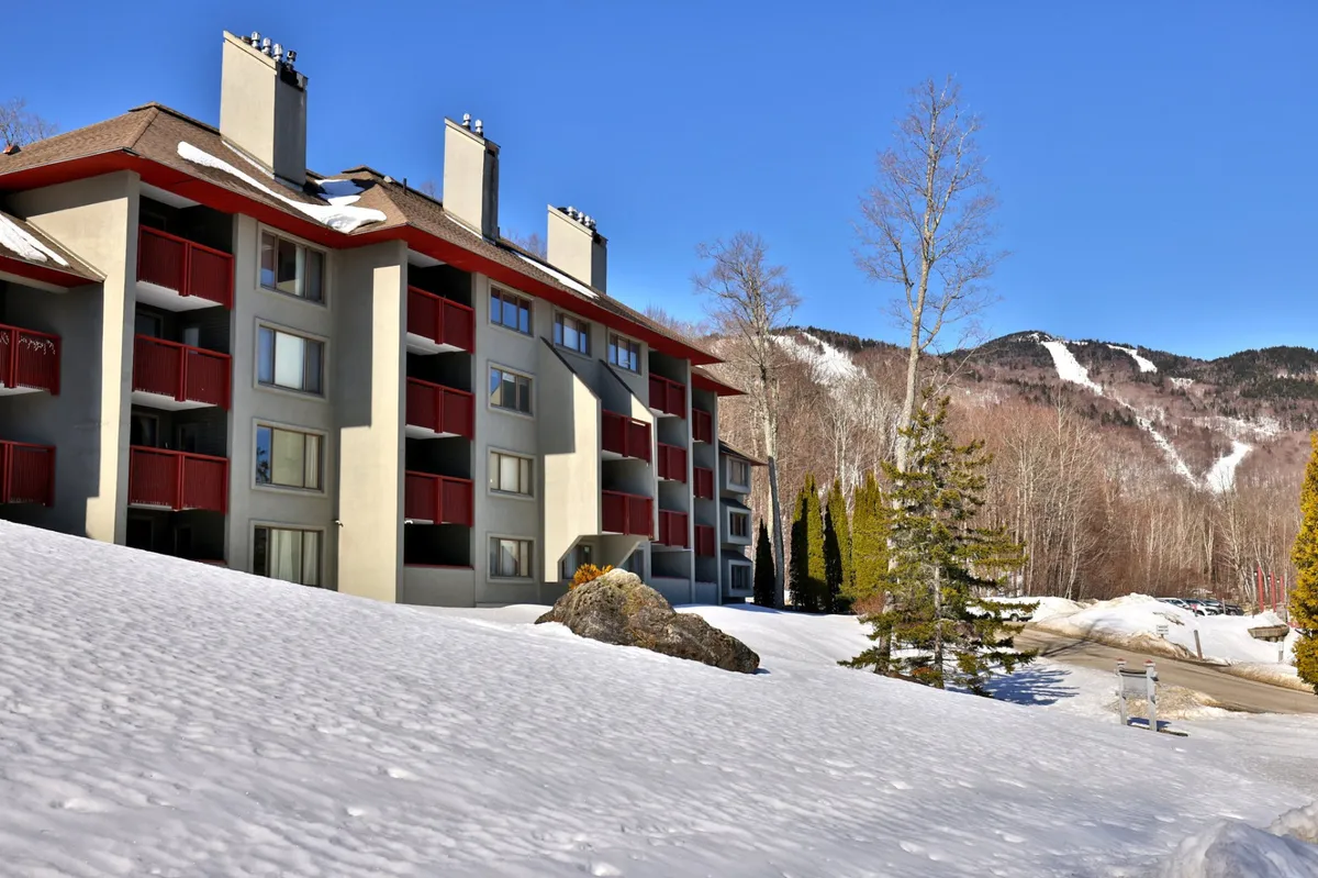 A scenic winter exterior with the building facade surrounded by snow, framed by trees and a dramatic mountain backdrop. The snow-covered ground and balcony emphasize a tranquil and natural setting, perfect for relaxation and wellness escapes.