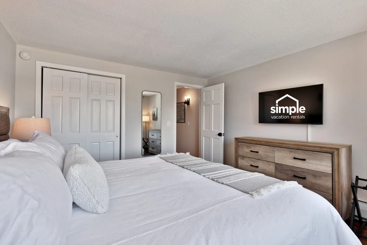 The photo showcases a king-sized bed with crisp white bedding, a wooden dresser, a wall-mounted TV, and a full-length mirror. Soft lighting and subtle mountain-inspired hues create a serene and inviting atmosphere, perfect for relaxation and restful sleep.