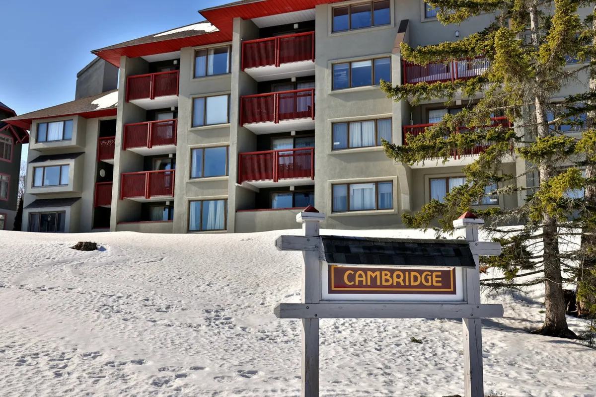 The exterior shot focuses on the Cambridge building sign, balconies, snow-covered ground, and surrounding trees. It conveys a welcoming base for adventure and relaxation, with immediate access to outdoor activities and inviting balconies.