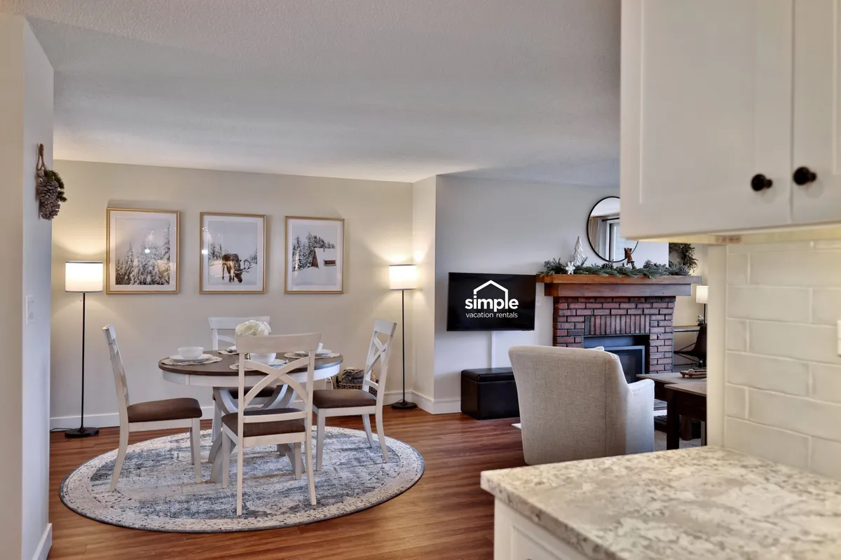 A dining area seamlessly integrated with a living space, featuring a large wooden dining table set near a glowing fireplace. Comfortable seating, wall art, and a television add to the welcoming, family-friendly atmosphere—ideal for shared meals, game nights, or planning adventures.