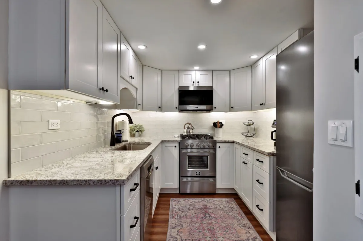 A spacious and modern kitchen featuring granite countertops, gleaming stainless steel appliances, white cabinetry, and a cozy rug underfoot. The ample counter space and inviting layout suggest plenty of room for meal preparation and socializing.