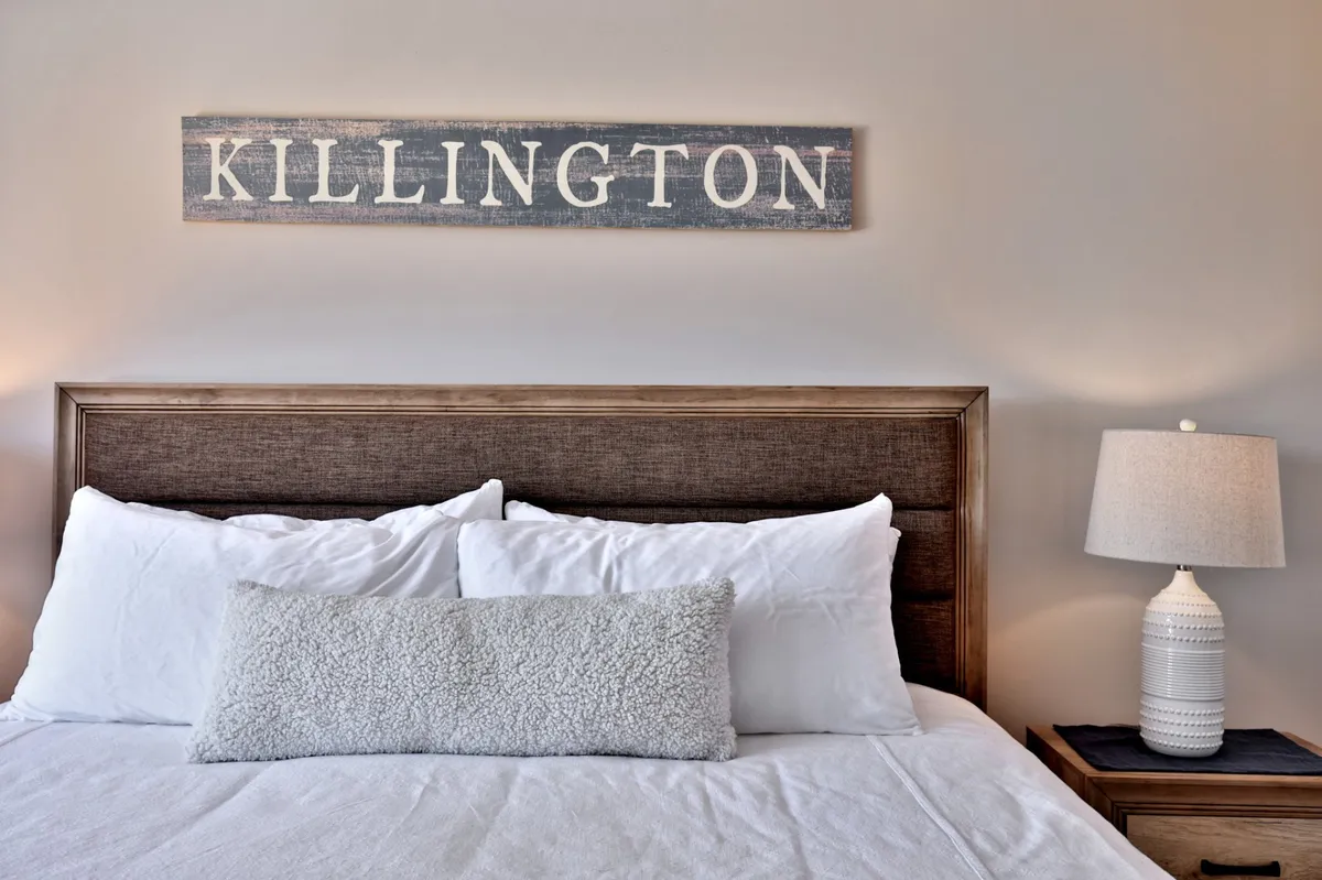 This close-up view features a textured headboard, decorative pillows, a softly glowing lamp, a wooden nightstand, and a sign reading 'KILLINGTON.' The scene exudes coziness and warmth, with an emphasis on soft lighting and elegant bedding.